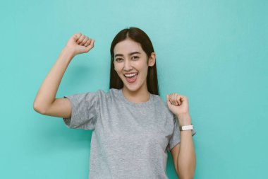Gri tişörtlü gülümseyen genç Asyalı kadın, kutlamada parlak turkuaz bir duvara karşı kollarını kaldırarak mutluluk, başarı ve pozitif enerjiyi temiz ve modern bir yaşam tarzıyla ifade ediyor..