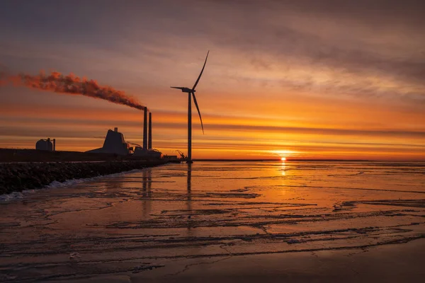 Oresund 'da enerji santrali ve kıyıya yakın modern bir yel değirmeni olan renkli bir gün doğumu. Su renkleri yansıtan buzla kaplıdır..