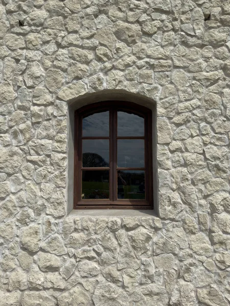 Geleneksel bir ahşap pencere desenli, pürüzlü taş bir duvara kurulmuş. Gökyüzü ve ağaçların yansımaları cam camlarda görünür..