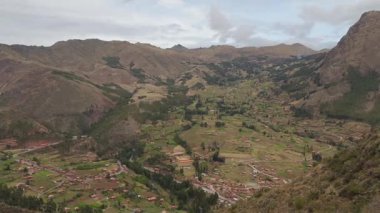 Geniş, bereketli İnkalar 'ın kutsal vadisini, teraslı tarlalar ve yuvarlanan yeşil ve kahverengi dağların arasına yerleştirilmiş küçük bir köyle geniş bir panoramik çekim yapar. Gökyüzü bulutlu ve bulutlu, dramatik manzarayı vurguluyor..