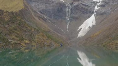 Laguna Humantay 'ın yavaş, yukarı doğru çekimleri, And Vadisi' nin yükseklerinde yer alan buzul gölü. Turkuaz su, karla kaplı, kayalık kayalık yüzeyi ve yukarıdaki sisli bir gökyüzünün altındaki dikey akıntıları yansıtır..