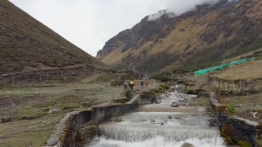 Bir grup yürüyüşçünün, And Vadisi 'nden dağlara doğru yürüyüşlerini geniş açıdan çekiyoruz. Önplanda beton taşmış bir yapısı olan berrak, kayalık bir dere akıyor. Etrafındaki yamaçlar dik ve kuru, tepemizde bulutlu bir gökyüzü var..