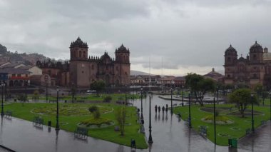 Cusco, Peru 'daki Plaza de Armas' ın gri, bulutlu ve yağmurlu bir günde çekilmiş yavaş ve dışa dönük bir çekimi. Meydanda yeşil çim ve lamba direkleri, Katedral ve İsa 'nın Şirketi Kilisesi var..