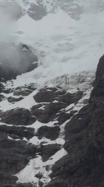 Dikey bir iz sürme çekimi, yüksek bir dağın engebeli, koyu kayalık yüzeylerinin beyaz kar parçalarıyla kaplı olduğunu ve sisli, gri bir gökyüzünün altında antik bir buzul olduğunu gösteriyor..