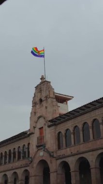 Cusco 'nun gökkuşağı renkli bayrağı gri, yağmurlu bir gökyüzünün altında kemerleri olan bir taş binanın tepesinde dalgalanıyor. Şehrin kalbindeki yerel gurur ve kimliğin sembolü..