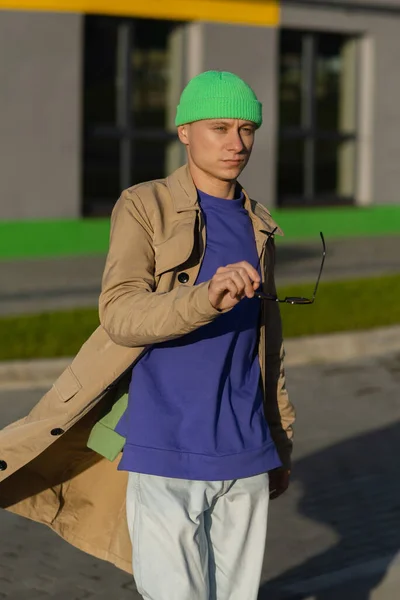 A young man wearing autumn clothes in the street. Young guy holding sunglasses and wearing coat, and green hat. — Stock Photo, Image