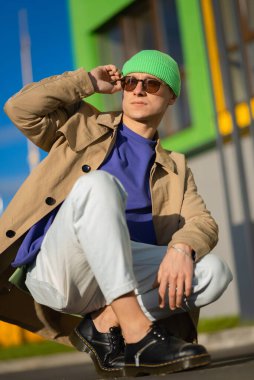 Modern bir binanın yanında şık yeşil şapkalı, şık kahverengi ceketli yakışıklı bir genç adam poz veriyor..