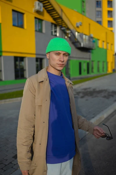 Atractivo hombre hipster urbano joven en ropa de moda de colores en una gorra de moda en gafas de sol pasea por la ciudad — Foto de Stock