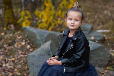 Mavi elbiseli, siyah ceketli, mavi gözlü bir sarışın, ağaçların arka planında, taşın üzerinde oturuyor. Doğayla uyum kavramı..