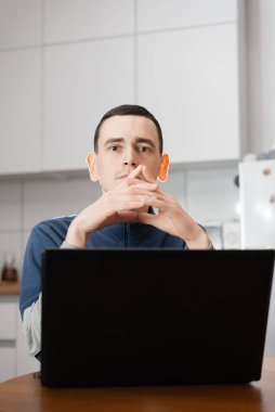 Sıradan giyimli genç girişimci yemek masasında oturuyor ve evinde dizüstü bilgisayarla çalışıyor.