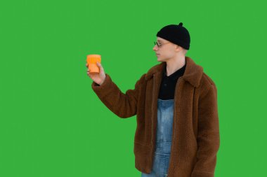 Güneş gözlüklü yakışıklı genç adam yeşil bir duvarın arkasında yeniden kullanılabilir bir bardak tutuyor.