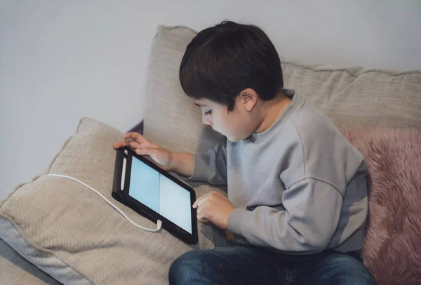 New normal life style,Kid sitting on sofa watching cartoons or playing game on tablet,Child boy using digital pad learning lesson on internet,Home schooling,Distance learning online education concept