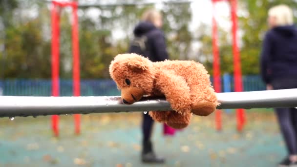 在雨天里 迷路的泰迪熊躺在秋千上 下着雨滴 孤独或悲伤的棕色熊独自躺下 模糊的孩子和父母在公园的背景下打秋千 — 图库视频影像