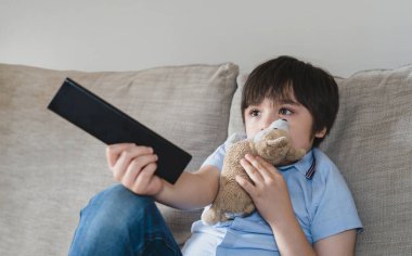 Tablet şarj olurken çizgi film izleyen mutlu çocuk, elinde uzaktan kumandayla kanepede oturan ve televizyon izleyen çocuk, okuldan sonra oturma odasında dinlenen çocuk. 