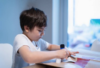 Anaokulu çocuğu okul ödevi için kağıda yapıştırıcı yapıştırıyor, küçük çocuk kağıda yapıştırıcı yapıştırıp DIY projesi yapıyor, çocuklar evde öğrenip oynuyor, ev okulunda oynuyor.