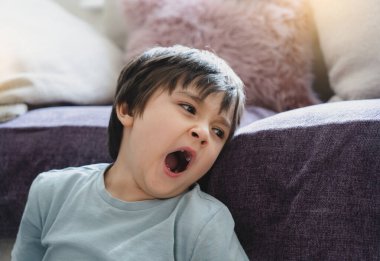 Otantik yorgun çocuk kanepenin yanında oturuyor, uykulu çocuk esniyor ve aşağı bakıyor, havanın değişmesi sırasında alerjisi olan çocuk, çocukluk dönemi toz maytundan ya da saman nezlesinden kaynaklanıyor, çocuklarda alerji var.