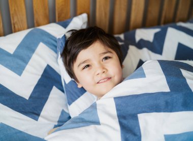 Yüzü kapalı bir şekilde kameraya gülümseyen çocuk gözleriyle bakan, büyük bir gülümsemeyle mutlu çocuğu çeken, seçici odaklı sağlıklı bir çocuk sabah yatakta dinlenen, pozitif çocuk konsepti.