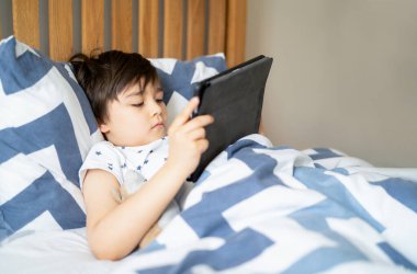 Portre okulundaki çocuk yatakta yatıp tablette hikaye okuyor, genç çocuk dijital ekranda oyun oynarken eğleniyor, mutlu çocuk sabahları yatak odasında dinleniyor, evde eğitim ya da online eğitim kavramını kullanıyor.