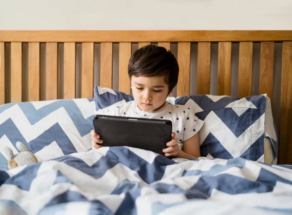Kid reading bed time stories on tablet before sleep, Happy boy sitting in bed playing games on digital pad, Child relaxing at home in his bed room on weekend.