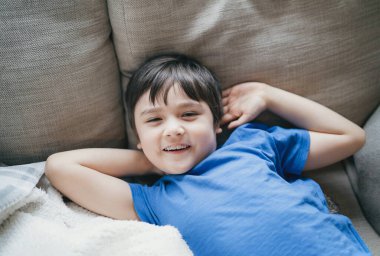 Kameraya gülümseyen çocuğun yüzünü kapat, hafta sonu koltukta uzanan mutlu bir çocuk, rahat bir çocuk kanepeye uzan ve pozitif çocuk konseptini izle.