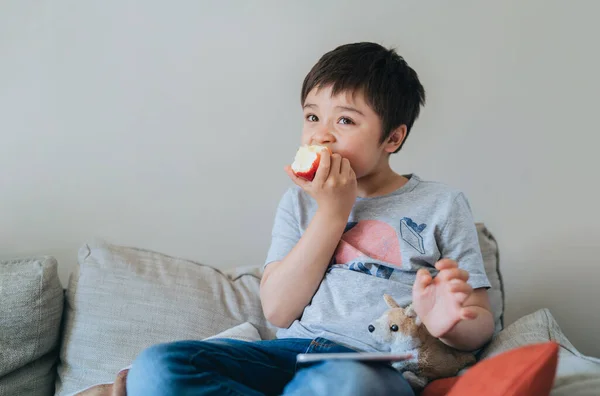 健康的な子供はテレビを見ながらリンゴを食べて 肖像画の子供は新鮮な果物を食べています 幸せな男の子座っているソファリラックスして自宅で週末 — ストック写真