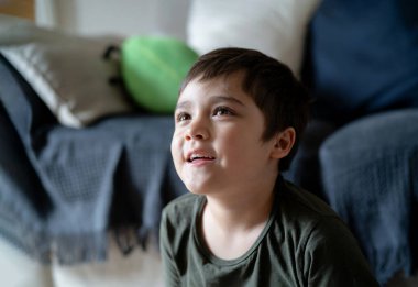 Mutlu çocuk gülümseyen yüzüyle yukarı bakıyor, pozitif küçük çocuk oturma odasında TV seyrediyor, sağlıklı çocuk evde eğleniyor, pozitif çocuk konsepti.