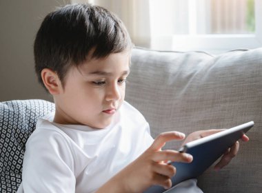 Portre çocuk kanepede uzanıp tablette çizgi film seyrediyor. Dokunmatik kâğıtta oyun oynayan tatlı çocuk. Oturma odasında tek başına eğlenen çocuk.