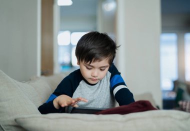 Çocuk kanepede oturup tablette çizgi film izlerken, çocuk dokunmatik kâğıtta oyun oynarken, çocuk oturma odasında kendi kendine eğlenirken, New normal yaşam tarzında.