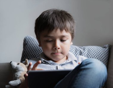 Portre çocuk kanepede oturmuş tablette çizgi film seyrediyor, şirin çocuk dokunmatik pedde oyun oynuyor, kapalı mekan portresi çekiyor çocuk çalışma odasında kendi kendine eğleniyor ve dinleniyor, yeni normal yaşam tarzı.