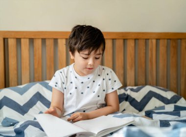 Portre Çocuk yatakta oturmuş kitap okuyor, genç çocuk yatak odasında kitap okuyor, gülen yüzlü mutlu çocuk sabah evde tek başına dinleniyor. Evde eğitim kavramı