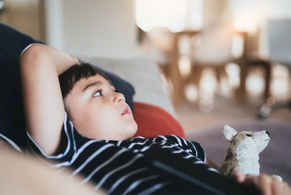 サイドビューの肖像画若い男の子のソファのテレビを見て 肯定的な子供のリビングルームで犬のおもちゃとソファに横たわって座っている 春か夏の週末に家でリラックスする子供 — ストック写真