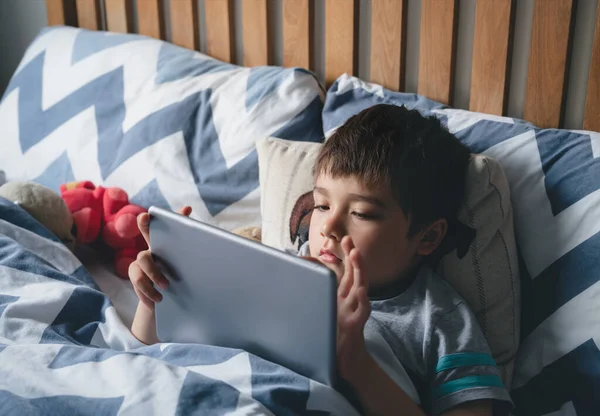Kid reading bed time stories on tablet before sleep, Happy boy sitting in bed playing games on digital pad, Child relaxing at home in his bed room on weekend.Children with technology concept