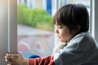 Happy kid sitting next to window playing with teddy bear, Cute boy playing alone, Positive child relaxing at home during cold weather outside in Autumn or Winter