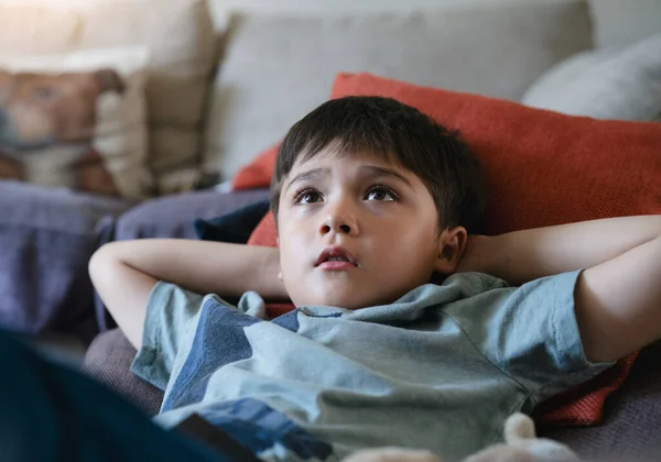 Happy boy lying on bean bag looking up watching cartoon on TV. mixed race Child resting in living room with bright light shining from window on sunny day summer. Kid relaxing at home on weekend
