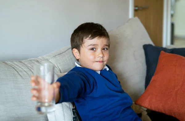 Happy school kid holding clear glass, Portrait mixed race child boy sitting on sofa showing empty glass of water after finished drinking.