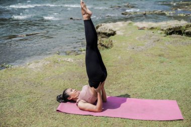 Zihinsel ve fiziksel sağlık. Sıska, bronzlaşmış, spor giyimli Asyalı bir kadın sabahın erken saatlerinde güzel bir nehir kıyısında yoga yapıyor. Kadın Sarvangasana ya da Omuz Durağı 'nı sahneliyor..