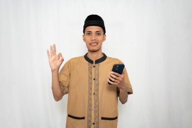 Geleneksel kıyafetleri olan genç bir adam elinde telefon tutup el sallıyor.