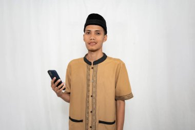 Geleneksel kıyafetler giyen genç bir adam stüdyo ortamında akıllı telefon tutuyor.