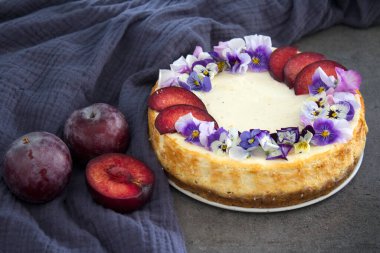 Kırmızı erikler ve yenilebilir çiçeklerle süslenmiş cheesecake. Kumaş dokusu arka planı. Ev yapımı tatlının en iyi görüntüsü.. 