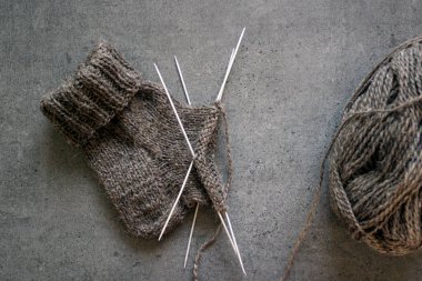 Çorap örüyorum, çalışıyorum. Yün ipliği, örgü iğneleri ve el yapımı çorabın yarısı. Gri desenli arka plan. Kış hobileri. 