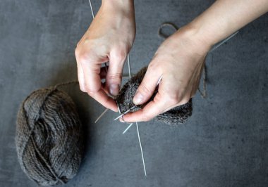 Kadın sıcak çorap örüyor. Örgü iğneleri, kahverengi çorap ve yün yumağı tutan ellerin yakın fotoğrafını çek. Gri desenli arka plan. Hobiler ve el sanatları konsepti. 