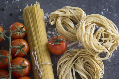 Masadaki makarna malzemeleri. Çiğ spagetti ve tagliatelle makarna, kiraz domatesleri ve arka plandaki gri dokuma üzerinde deniz tuzu fotokopisi. Sağlıklı yeme kavramı.