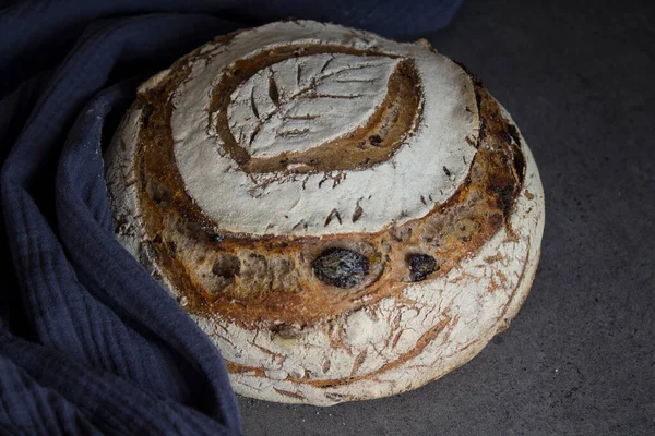 Sourdough bread with a leaf pattern on the top. Whole grain bread with black olives, raisins and nuts. Healthy eating concept. Long-fermented sourdough loaf top view photo. Beautiful round whole grain bread on a table. Still life food.