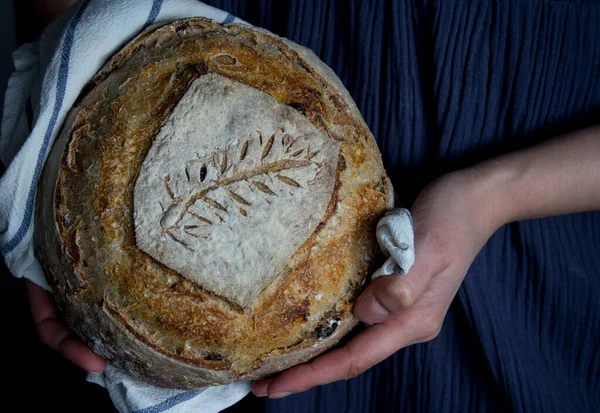 Close up photo of woman\'s hands holding fresh artisan bread with wheat pattern on the top. Beautifully scored round sourdough bread. Healthy eating concept.