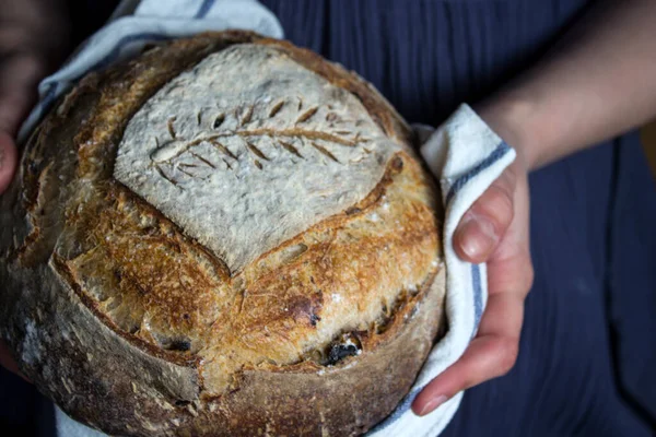 Close up photo of woman's hands holding fresh artisan bread with wheat pattern on the top. Beautifully scored round sourdough bread. Healthy eating concept.