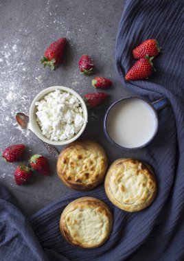 Vatrushka - Geleneksel Rus turtası ve süzme peynir. Tatlı çörekler, süzme peynir, sütlü mavi seramik bardak, sulu çilekler ve lacivert mutfak havlusu. Gri dokulu arkaplan.