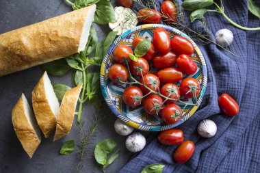 Brüksel usulü taze malzemeler. Mavi seramik tabakta sulu kiraz domatesleri fesleğen yaprakları, sarımsak soğanları baget ekmeği ve lacivert mutfak havlusunda otlar. Gri desenli arka plan. Otantik İtalyan tarifleri.