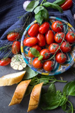 Brüksel usulü taze malzemeler. Mavi seramik tabakta sulu kiraz domatesleri fesleğen yaprakları, sarımsak soğanları baget ekmeği ve lacivert mutfak havlusunda otlar. Gri desenli arka plan. Otantik İtalyan tarifleri.