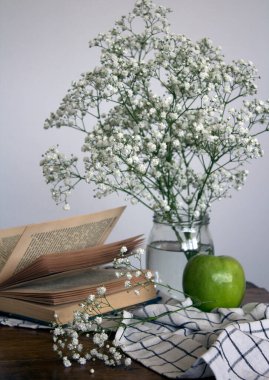Küçük beyaz çiçekli cam kavanoz, yeşil elma ve açık bir kitap. Basit kompozisyon, beyaz arka planda çingene çiçekleri ile yaşam devam ediyor.. 