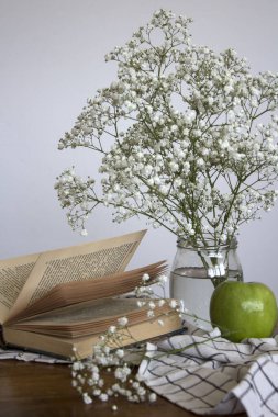 Küçük beyaz çiçekli cam kavanoz, yeşil elma ve açık bir kitap. Basit kompozisyon, beyaz arka planda çingene çiçekleri ile yaşam devam ediyor.. 